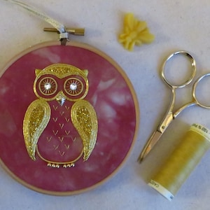 May include: A gold embroidered owl on a red fabric background in a wooden embroidery hoop. The owl has sparkling eyes and is surrounded by gold thread. There are sewing scissors, a spool of gold thread, and a piece of beeswax on the table.