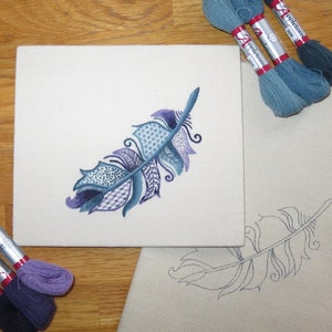 May include: A close-up of a hand-stitched embroidery project featuring a blue and purple feather design on a white canvas. The embroidery is surrounded by spools of blue and purple embroidery floss.
