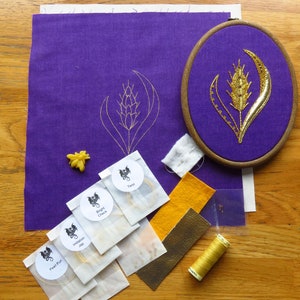 May include: A purple fabric with a gold embroidered wheat stalk design in a wooden embroidery hoop. There are several small white envelopes with gold thread and a small yellow wax seal.