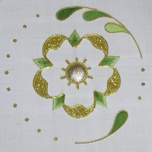 May include: A white fabric with a gold and green embroidered flower design. The flower has a silver centre and is surrounded by green leaves.