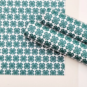 May include: White and green patterned fabric featuring a repeating clover design with the letter "H" inside each clover. The fabric is shown in a flat sheet and rolled up, showcasing the pattern's continuity.