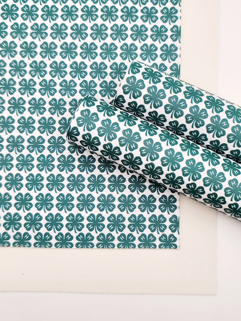 May include: White and green patterned fabric featuring a repeating clover design with the letter "H" inside each clover. The fabric is shown in a flat sheet and rolled up, showcasing the pattern's continuity.