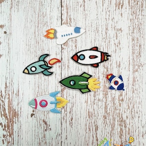 May include: Six colorful embroidered iron-on patches featuring rockets and a spaceship. The patches are on a light wood background.