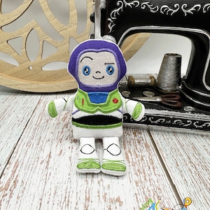 Puede incluir: Juguete de peluche bordado de Buzz Lightyear de la franquicia Toy Story. El juguete es blanco con detalles verdes y negros. El juguete tiene un casco morado y una cara amigable.
