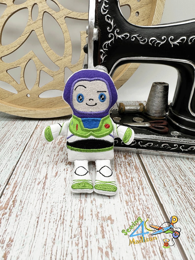 Puede incluir: Juguete de peluche bordado de Buzz Lightyear de la franquicia Toy Story. El juguete es blanco con detalles verdes y negros. El juguete tiene un casco morado y una cara amigable.