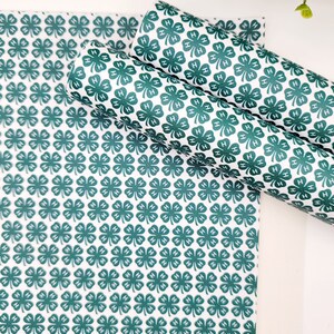 May include: White and green patterned fabric featuring a repeating clover design with the letter "H" inside each clover. The fabric is shown in a flat sheet and rolled into two cylinders.