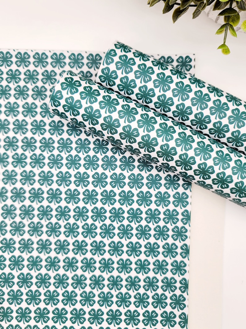 May include: White and green patterned fabric featuring a repeating clover design with the letter "H" inside each clover. The fabric is shown in a flat sheet and rolled into two cylinders.