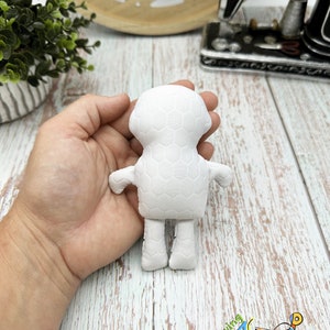 Puede incluir: Mu&ntilde;eca de tela blanca con un patr&oacute;n de panal. La mu&ntilde;eca tiene una cabeza redonda, dos brazos, dos piernas y un cuerpo peque&ntilde;o. La mu&ntilde;eca est&aacute; sostenida en la mano de una persona.
