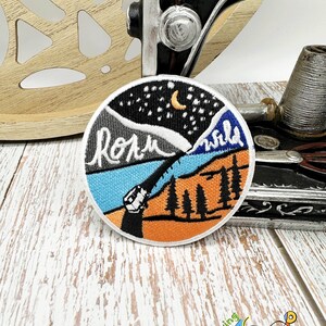 May include: A round embroidered patch with a night sky, mountains, and trees. The patch has the words "roam wild" stitched in white thread.