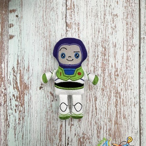 Puede incluir: Un juguete de peluche de Buzz Lightyear de la franquicia Toy Story. El juguete es blanco con detalles verdes y negros. El juguete tiene un casco morado con una cara sonriente y un solo ojo.