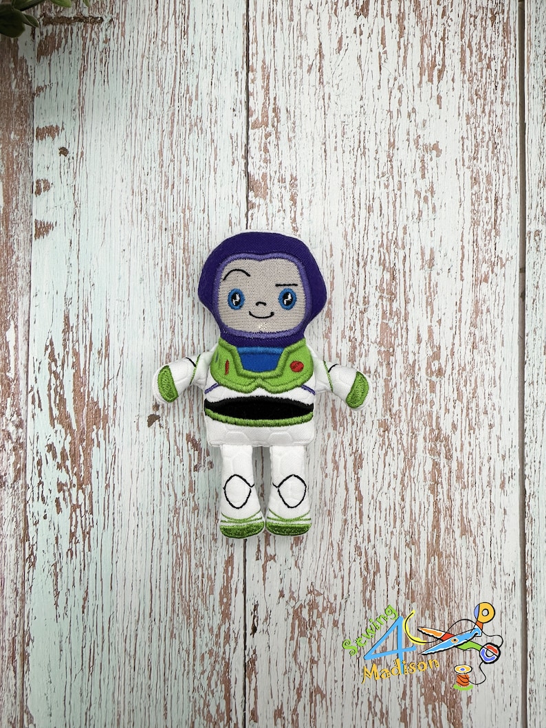Puede incluir: Un juguete de peluche de Buzz Lightyear de la franquicia Toy Story. El juguete es blanco con detalles verdes y negros. El juguete tiene un casco morado con una cara sonriente y un solo ojo.