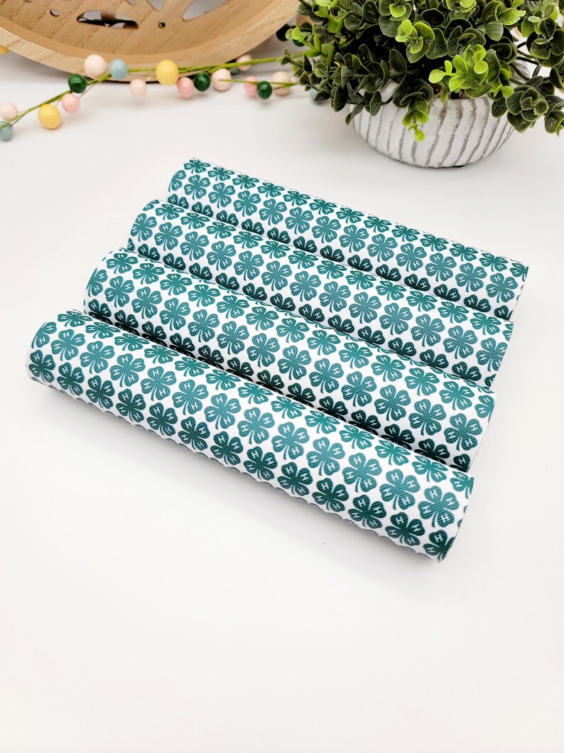 May include: Four rolls of white fabric with a repeating pattern of green four-leaf clovers. The clovers have the letters "H" inside each leaf. The fabric is rolled up and stacked on top of each other.