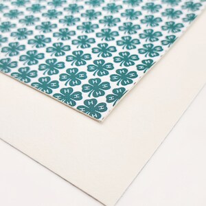 May include: A close-up of two pieces of material. The top piece has a white background with a repeating pattern of teal four-leaf clovers, each with the letter "H". The bottom piece is a solid off-white color.