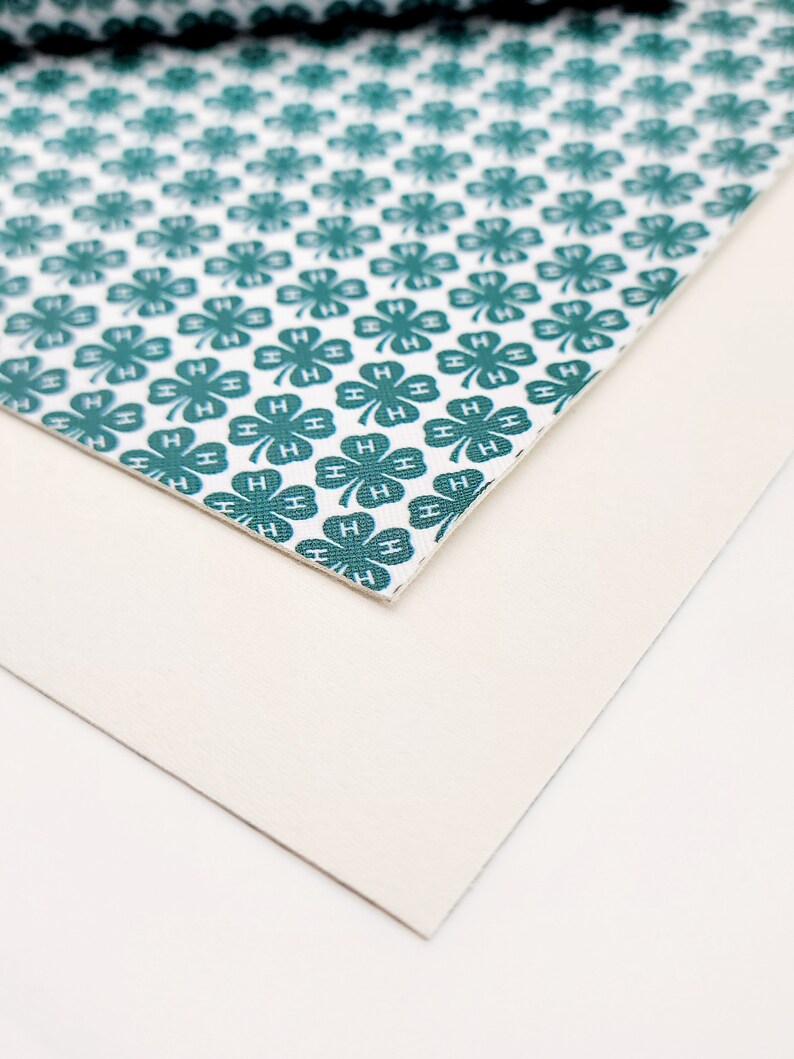May include: A close-up of two pieces of material. The top piece has a white background with a repeating pattern of teal four-leaf clovers, each with the letter "H". The bottom piece is a solid off-white color.