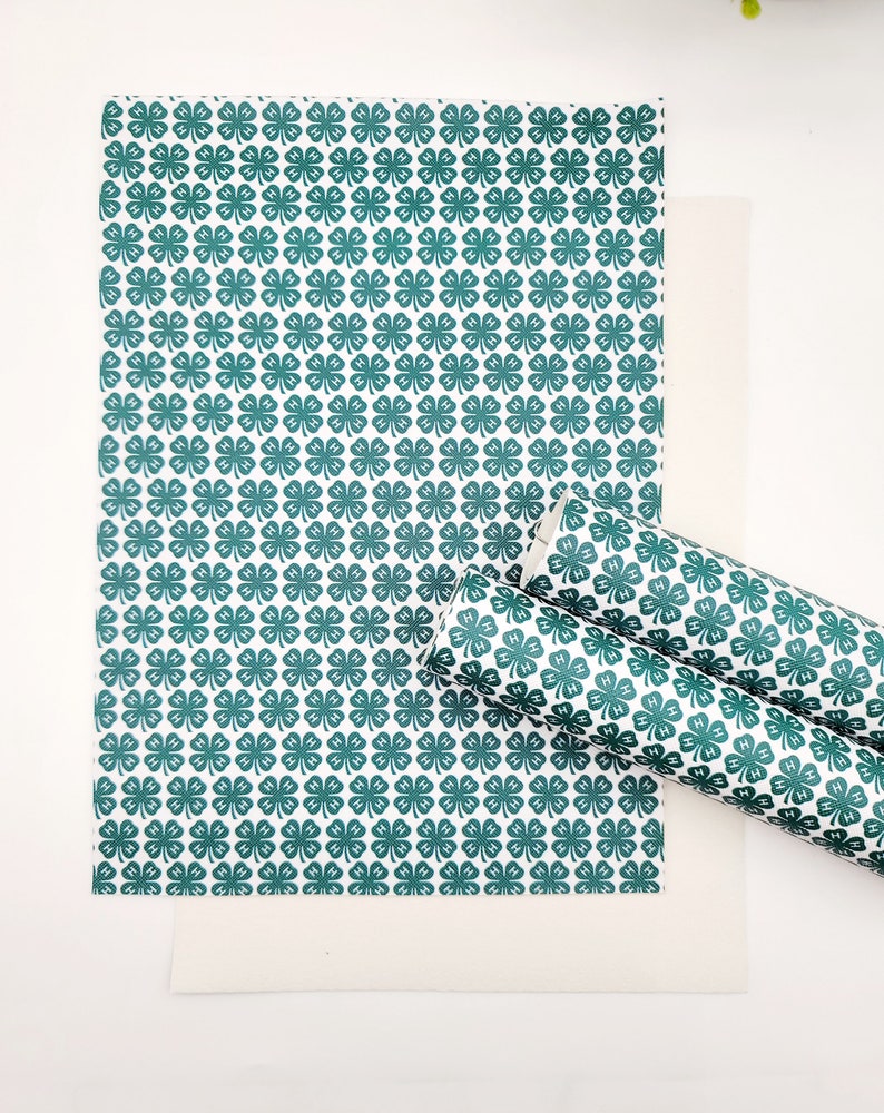 May include: White background with a sheet and a rolled piece of paper featuring a repeating pattern of green four-leaf clovers. The clover design is a dark teal color.