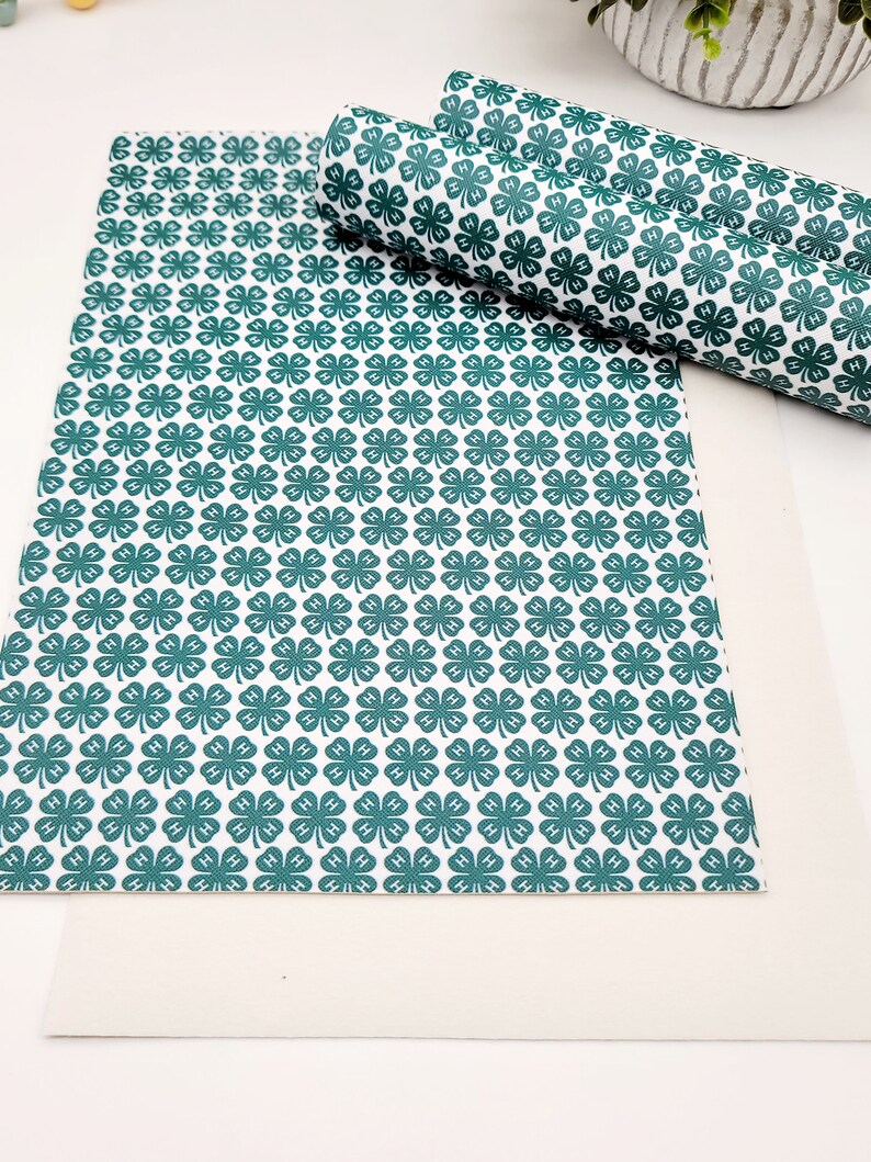 May include: White and teal clover patterned faux leather sheets, one flat and two rolled. The clover design is repeated across the surface. The material is suitable for crafting projects, such as making bows, earrings, or other accessories.