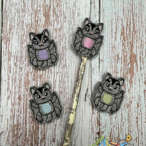 May include: Four gray felt pencil toppers shaped like flying squirrels with different colored accents. The toppers are on a wooden surface with a pencil in the center.