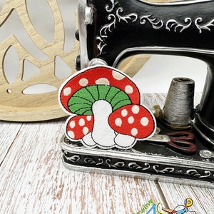 May include: A red and white mushroom with green accents is embroidered on a black sewing machine. The machine is on a wooden surface.