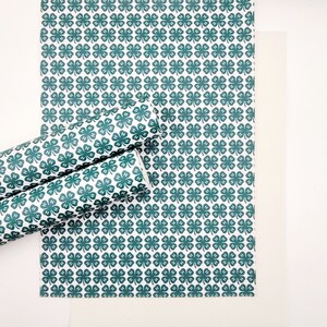 May include: White and green patterned paper with a repeating clover design. The image shows a flat sheet of paper, a rolled-up section, and a plain white sheet. The clover pattern includes the letters "H" in each clover.