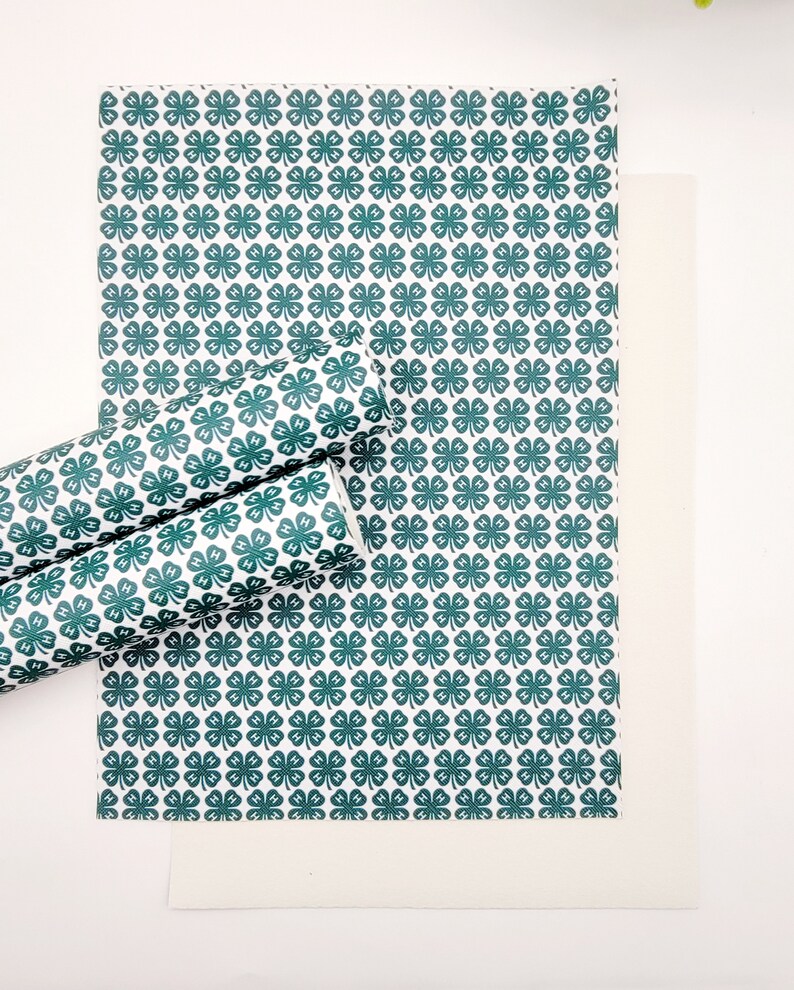 May include: White and green patterned paper with a repeating clover design. The image shows a flat sheet of paper, a rolled-up section, and a plain white sheet. The clover pattern includes the letters "H" in each clover.