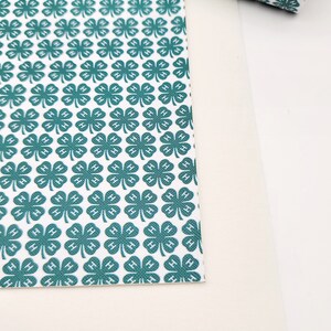 May include: White and teal green clover patterned fabric. The fabric features a repeating pattern of four-leaf clovers with the letter "H" in each leaf. A rolled-up piece of the fabric is visible in the background.