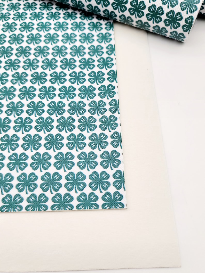May include: White and teal green clover patterned fabric. The fabric features a repeating pattern of four-leaf clovers with the letter "H" in each leaf. A rolled-up piece of the fabric is visible in the background.