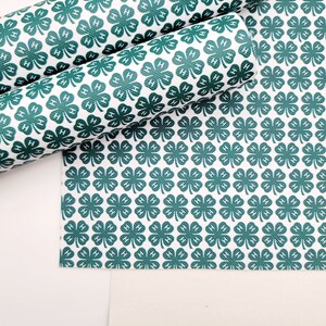 May include: White material with a repeating pattern of dark green four-leaf clovers, each with an "H" in the center. Some of the material is rolled up, while other pieces are flat. The background is white.