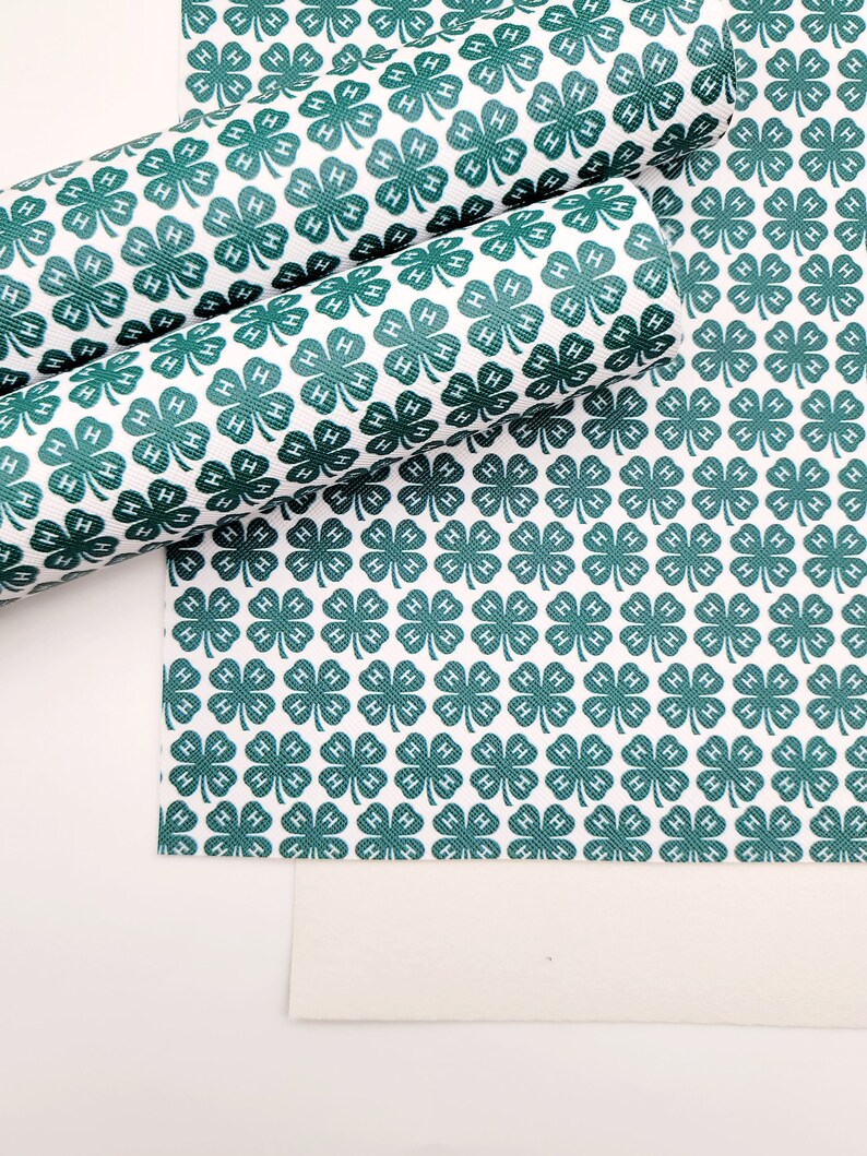 May include: White material with a repeating pattern of dark green four-leaf clovers, each with an "H" in the center. Some of the material is rolled up, while other pieces are flat. The background is white.