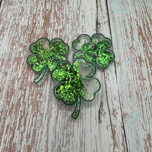 May include: Three green glitter shamrock charms with a clear resin background. The charms are on a white wooden surface.