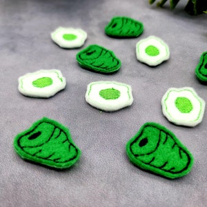 May include: Nine felt patches, six white and three green, with embroidered images of eggs and steaks. The patches are on a gray surface.
