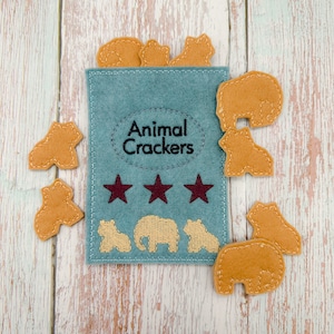 May include: A blue felt pouch with the words "Animal Crackers" embroidered on it. Three gold stars are above the text. The pouch is surrounded by felt animal crackers in various shapes, including elephants, bears, and hippos.