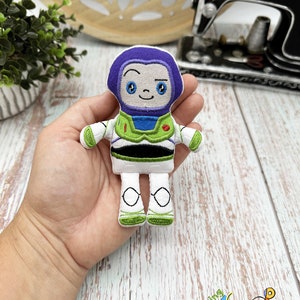 May include: A small, embroidered plush toy of Buzz Lightyear from the Toy Story franchise. The toy is white with green, blue, and purple accents. It has a friendly face with big eyes and a smile.