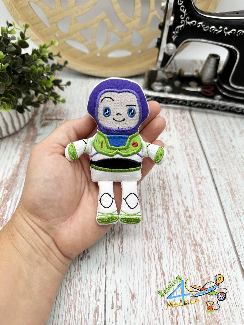 Puede incluir: Un peque&ntilde;o juguete de peluche bordado de Buzz Lightyear de la franquicia Toy Story. El juguete es blanco con detalles verdes, azules y morados. Tiene una cara amigable con ojos grandes y una sonrisa.