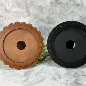 May include: Two decorative wooden coasters. One is light brown with a scalloped edge, and the other is black with a scalloped edge. Both have a circular centre hole. The coasters are on a white surface with greenery.