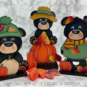 May include: Three decorative black bear figures with autumn themes. One wears a green hat with patches, one sits on a pumpkin wearing a yellow hat, and one wears a green sweater and scarf. Each bear is accented with pumpkins and leaves. Text reads: AVAILABLE AS A SET OF 3 BEARS OR INDIVIDUAL.
