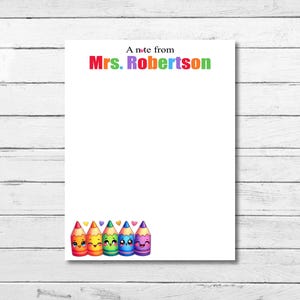 Peut inclure: Bloc-notes blanc avec le texte "A note from Mrs. Robertson" en lettres colorées. Le bas du bloc-notes présente une rangée de crayons de couleur souriants de style dessin animé. Le fond est une surface en bois blanc.