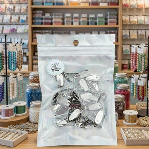 May include: A transparent bag containing numerous silver metal clasps. The bag features a sticker with the text "Loop & Loop by Katie". The background displays a craft shop with various beads and jewellery-making materials.