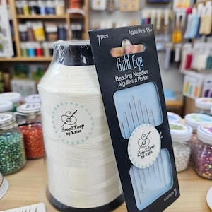 May include: A large spool of white thread and a packet of Gold Eye beading needles. The thread spool has a Loop2Loop by Katie sticker. The needle packet is black with the text "Gold Eye Beading Needles" and "Ages/Ans 15+".