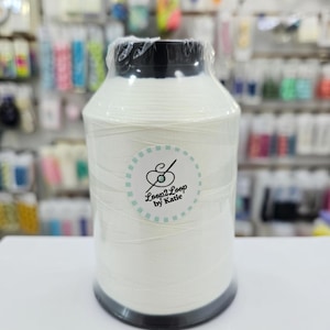 May include: A large spool of white thread, wrapped in clear plastic, with a black cap and base. A circular sticker on the spool reads "Loop2Loop by Katie" in a teal and white design. The background is a blurred display of craft supplies.