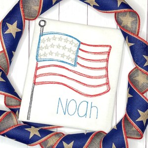 May include: White embroidered t-shirt with a red, white, and blue American flag and the name "Noah" in blue thread.