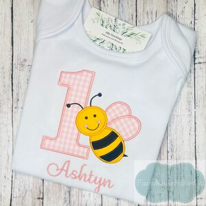 May include: White onesie with a pink and white gingham number one and a yellow and black embroidered bee. The onesie is personalized with the name "Ashtyn" in pink.