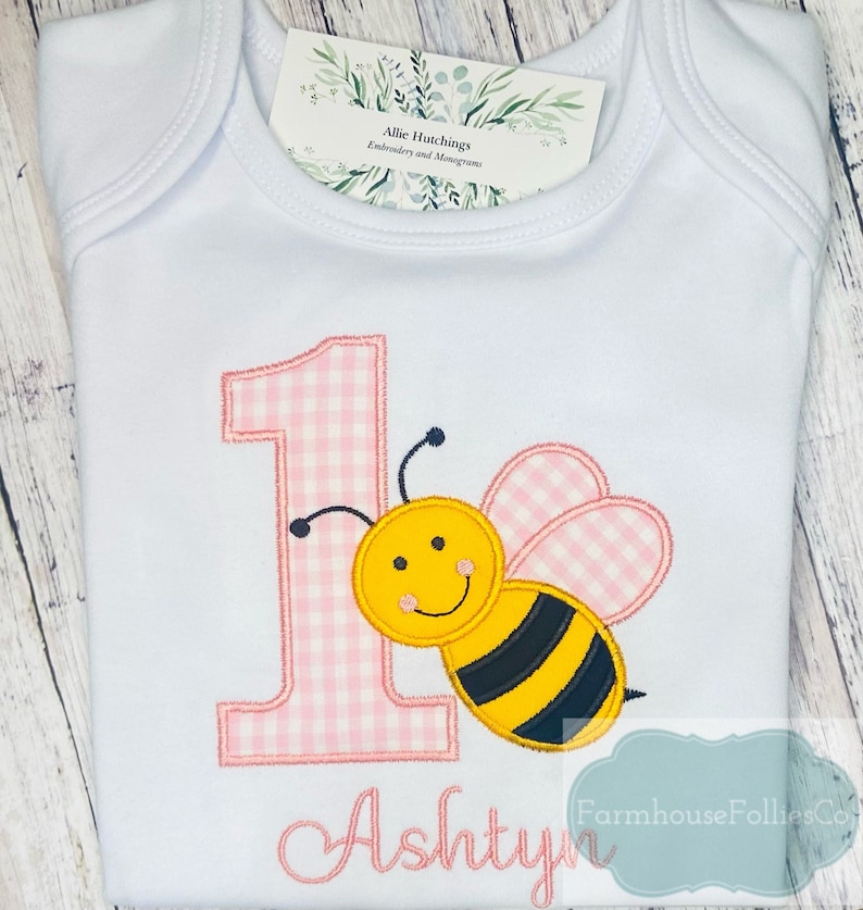 May include: A white onesie with pink and white gingham fabric embroidered with the number "1" and a yellow and black bee. The name "Ashtyn" is embroidered below the bee.