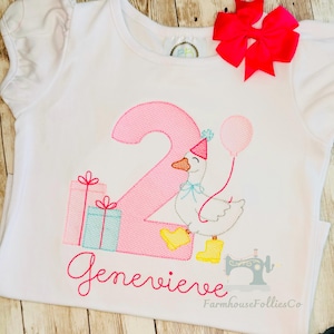 May include: A white short-sleeved t-shirt featuring an embroidered design of a large pink number two, a white duck wearing a pink party hat and yellow boots, two gift boxes, a pink balloon, and the name Genevieve in pink cursive script.