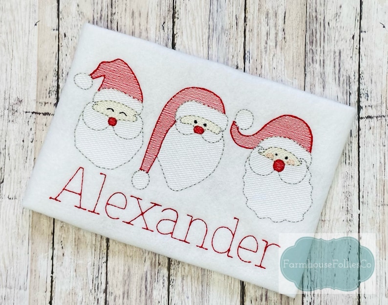 K&ouml;nnte beinhalten: Wei&szlig;er bestickter Weihnachtsstrumpf mit drei roten Weihnachtsmann-Gesichtern und dem Namen "Alexander" in roten Buchstaben.