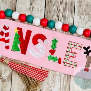 Könnte beinhalten: Rosa rechteckiges Schild mit dem Namen "ANSLEY" in festlichen Buchstaben zum Thema Weihnachten bestickt. Die Buchstaben sind mit Zuckerstangenstreifen, einem Weihnachtsbaum, einem Weihnachtsmannhut, Stechpalmen und einem Rentier verziert. Eine Perlenkette umgibt das Schild.