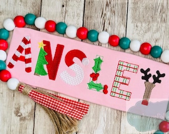 Könnte beinhalten: Rosa rechteckiges Schild mit dem Namen "ANSLEY" in festlichen Buchstaben zum Thema Weihnachten bestickt. Die Buchstaben sind mit Zuckerstangenstreifen, einem Weihnachtsbaum, einem Weihnachtsmannhut, Stechpalmen und einem Rentier verziert. Eine Perlenkette umgibt das Schild.