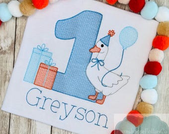 Könnte beinhalten: Weißes besticktes Shirt mit einer großen blauen Zahl 1, einer Cartoon-Gans mit einem Ballon und zwei Geschenkboxen. Der Name "Greyson" ist darunter gestickt. Das Shirt ist mit einer Girlande aus bunten Pompons verziert.