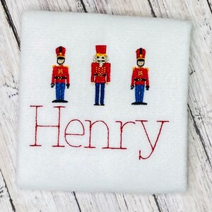 May include: White fabric with three red, blue, and yellow toy soldier figures and the name "Henry" embroidered in red.