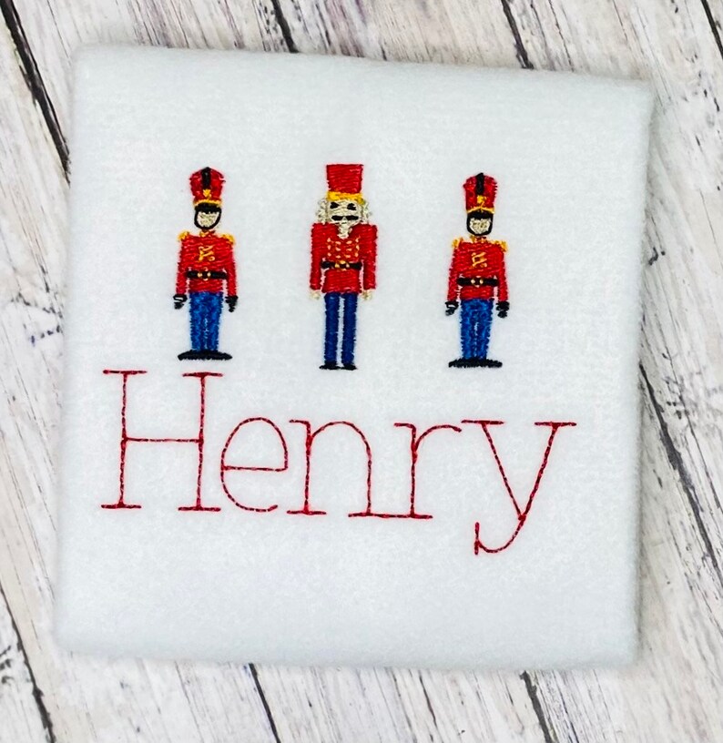 May include: White fabric with three red, blue, and yellow toy soldier figures and the name "Henry" embroidered in red.