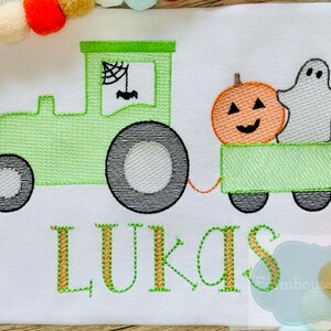 Op de afbeelding: Een groene tractor trekt een wagen met een pompoen en een spook. De wagen is groen en heeft zwarte wielen. De tractor heeft een spinnenweb in het raam. De naam "LUKAS" is in oranje en groen geborduurd aan de onderkant van de afbeelding.